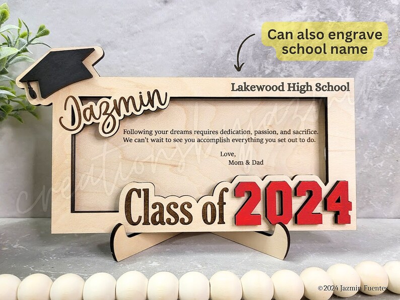 May include: Wooden graduation photo frame with the name "Dazmin" and "Class of 2024" in red. The frame includes the text "Lakewood High School" and a graduation cap. The frame has a stand and includes a message from Mom & Dad.