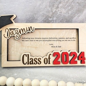 May include: A wooden graduation photo frame with the name "Jazmin" and "Class of 2024" in red. The frame includes a graduation cap and a message from Mom & Dad.