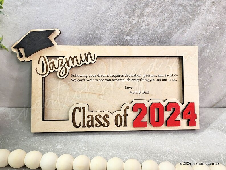 May include: A wooden graduation photo frame with the name "Jazmin" and "Class of 2024" in red. The frame includes a graduation cap and a message from Mom & Dad.