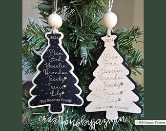 May include: Two wooden Christmas tree ornaments. One is black with white trim, the other is natural wood. Both have family names and the year 2024. The ornaments are hanging on a Christmas tree.