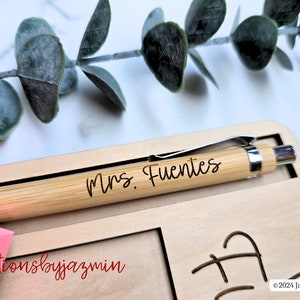 May include: A personalised bamboo pen, engraved with "Mrs. Fuentes", lies on a wooden desk organiser. The organiser features a pen slot, space for sticky notes, and decorative cutouts. A pink sticky note pad and eucalyptus leaves are also visible in the image.