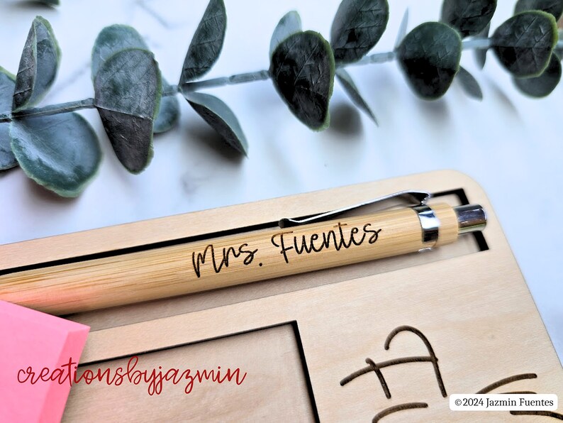 May include: A personalised bamboo pen, engraved with "Mrs. Fuentes", lies on a wooden desk organiser. The organiser features a pen slot, space for sticky notes, and decorative cutouts. A pink sticky note pad and eucalyptus leaves are also visible in the image.
