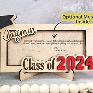 May include: A wooden graduation frame with the name "Dazmin" and "Class of 2024" in red. The frame includes a graduation cap and a message from "Mom & Dad". The frame is designed to hold a photo and has a stand.