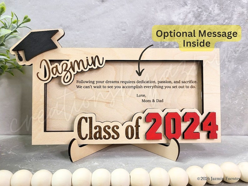 May include: A wooden graduation frame with the name "Dazmin" and "Class of 2024" in red. The frame includes a graduation cap and a message from "Mom & Dad". The frame is designed to hold a photo and has a stand.