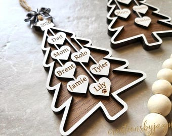 May include: A wooden Christmas tree ornament featuring a dark brown wood center and a white border. It includes a black and white buffalo plaid bow at the top with the year 2026. Several white heart-shaped wooden pieces are attached, engraved with the names Dad, Mom, Rolo, Brent, Tyler, Jamie, and Lily, with a paw print icon next to Lily.