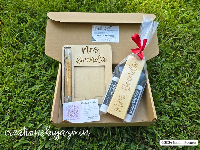 May include: A gift box containing a wooden picture frame, pen, and bookmark, all engraved with "Mrs. Brenda." The box also includes two Expo dry-erase markers. The items are arranged on a bed of grass.
