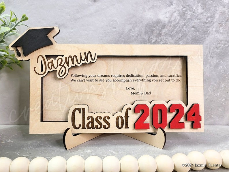 May include: A wooden graduation frame with the name "Dazmin" and "Class of 2024" in a decorative font. The frame includes a graduation cap and a message from Mom & Dad. The frame is designed to hold a photo.