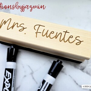 May include: A light-coloured wooden dry erase board eraser engraved with the name "Mrs. Fuentes". Two black and white Expo dry erase markers are in the foreground. The text "creationsbyjazmin" is in the top left corner.