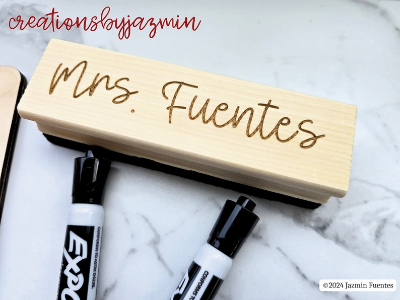 May include: A light-coloured wooden dry erase board eraser engraved with the name "Mrs. Fuentes". Two black and white Expo dry erase markers are in the foreground. The text "creationsbyjazmin" is in the top left corner.