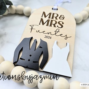 May include: Wooden wedding ornament with "MR & MRS Fuentes 2024" text. Features a black tuxedo and white dress cutout design. Includes a white ribbon for hanging. The ornament is part of a decorative bead garland.