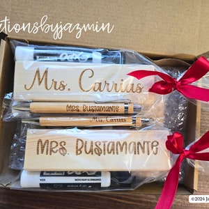 May include: A gift box containing personalized wooden desk nameplates and pens. The nameplates are engraved with "Mrs. Carrias" and "Mrs. Bustamante". The pens are bamboo with engraved names. The box is tied with red ribbons.