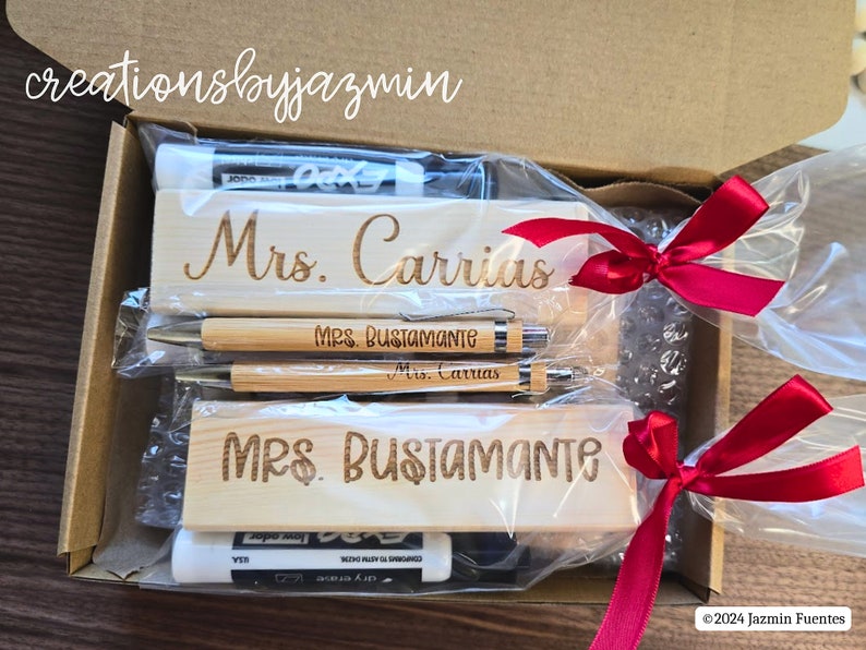 May include: A gift box containing personalized wooden desk nameplates and pens. The nameplates are engraved with "Mrs. Carrias" and "Mrs. Bustamante". The pens are bamboo with engraved names. The box is tied with red ribbons.