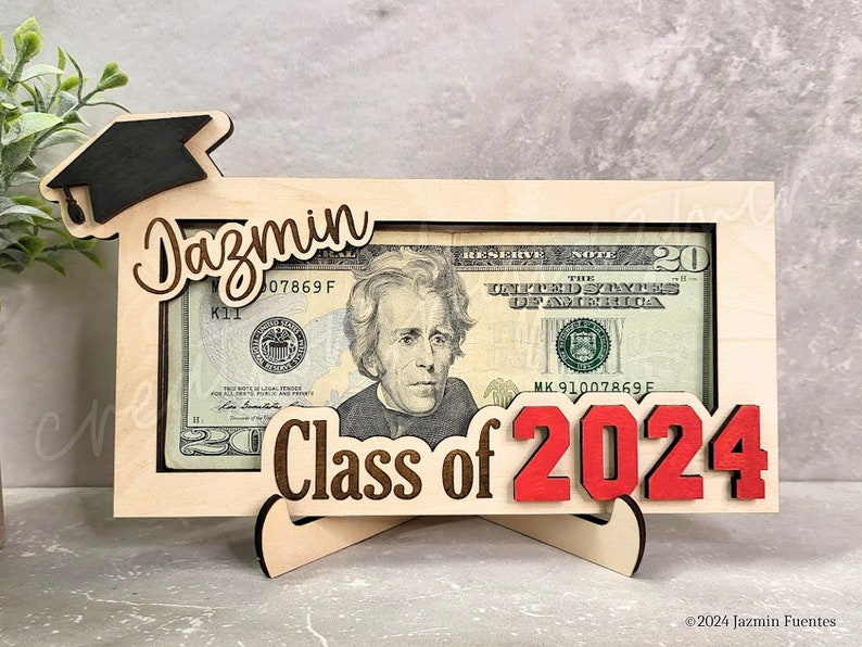 May include: A wooden graduation money holder with a $20 bill visible. The frame features the name "Jazmin", the words "Class of 2024", and a graduation cap. The numbers "2024" are in red.