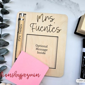 May include: A wooden desk organiser engraved with "Mrs. Fuentes". It features a pen, a pink sticky note, and a frame for a message. A black dry-erase marker and eraser are also visible.
