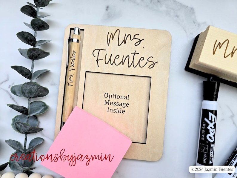 May include: A wooden desk organiser engraved with "Mrs. Fuentes". It features a pen, a pink sticky note, and a frame for a message. A black dry-erase marker and eraser are also visible.