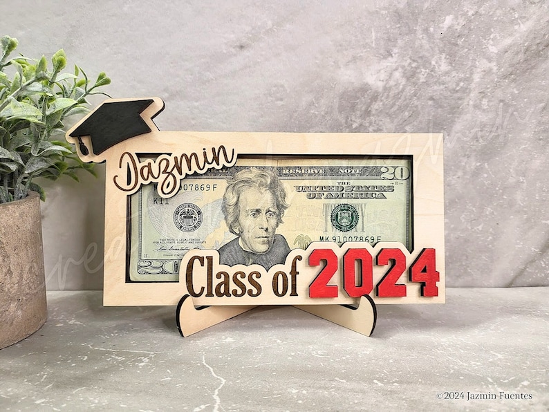 May include: A wooden graduation money holder with the name "Jazmin" and "Class of 2024" in red. The frame holds a twenty-dollar bill. A black graduation cap is attached to the top left corner. The holder is supported by a wooden stand.