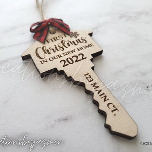 May include: Wooden key-shaped Christmas ornament with the text "First Christmas in our New Home 2022" and "123 Main Ct." A red and black plaid bow is attached to the top. The ornament is made of light-colored wood.