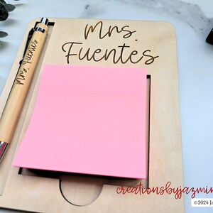 May include: A wooden desk organiser with a pink sticky note pad and a pen. The pen and the organiser are engraved with the name "Mrs. Fuentes". The organiser also has the text "creationsbyjazmin" and "&copy; 2024 Jazmin Fuentes".