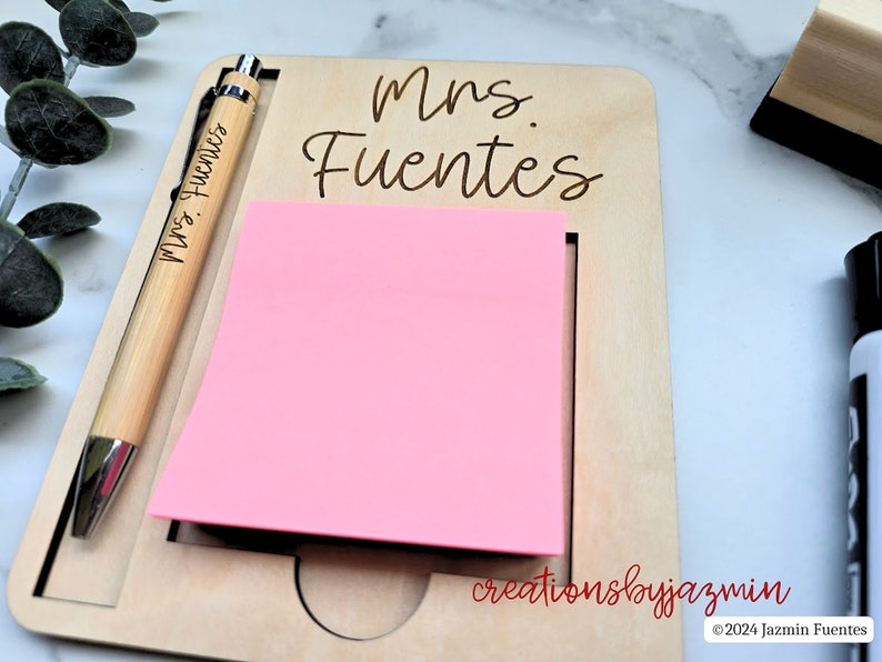 May include: A wooden desk organiser with a pink sticky note pad and a pen. The pen and the organiser are engraved with the name "Mrs. Fuentes". The organiser also has the text "creationsbyjazmin" and "&copy; 2024 Jazmin Fuentes".