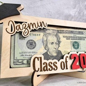 May include: A wooden graduation-themed money holder with a black mortarboard, the name "Dazmin", and "Class of 2024" in red. The frame holds a twenty-dollar bill. The holder is supported by a wooden stand.