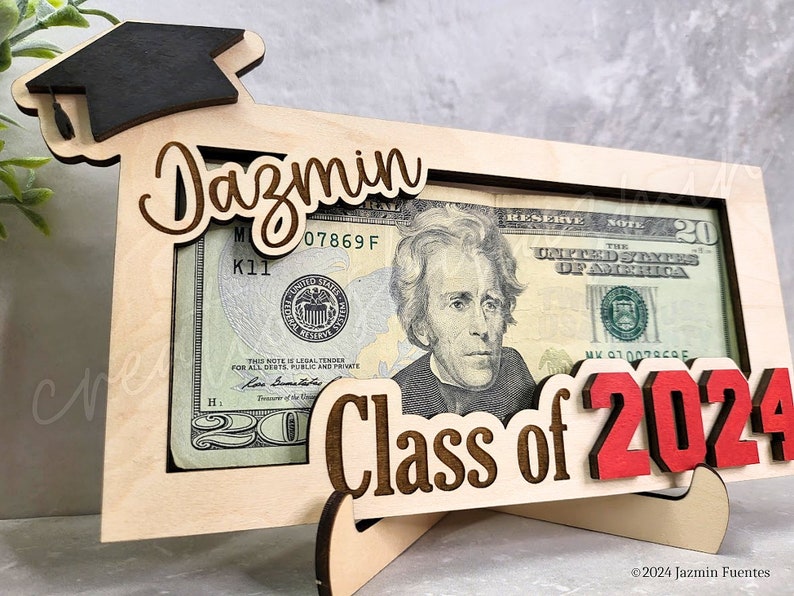 May include: A wooden graduation-themed money holder with a black mortarboard, the name "Dazmin", and "Class of 2024" in red. The frame holds a twenty-dollar bill. The holder is supported by a wooden stand.