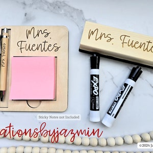 May include: A personalised wooden desk organiser set with the name "Mrs. Fuentes". The set includes a notepad holder with a pen, a whiteboard eraser, and two black dry-erase markers. The notepad holder has a pink sticky note pad.