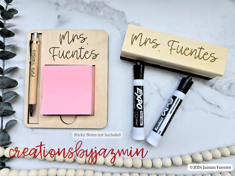 May include: A personalised wooden desk organiser set with the name "Mrs. Fuentes". The set includes a notepad holder with a pen, a whiteboard eraser, and two black dry-erase markers. The notepad holder has a pink sticky note pad.