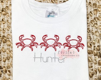 Könnte beinhalten: Weißes T-Shirt mit drei roten gestickten Krabben und dem Namen "Hunter" in einer grauen Stickerei. Das Shirt hat ein kleines weißes Etikett mit dem Text "GRB Blanks" darauf. Das Shirt liegt auf einem gewebten braunen Untergrund.