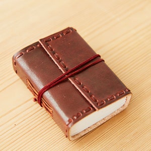 Op de afbeelding: Een klein, bruin, lederen gebonden dagboek met een rode koordsluiting. Het dagboek heeft decoratieve stiksels rond de randen.