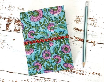 May include: A blue and pink floral patterned notebook with a red and brown string tied around it. The notebook is lying on a white wooden surface with a pencil next to it.