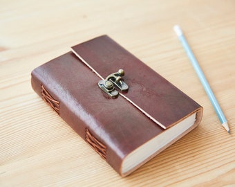 May include: A brown leather-bound journal with a brass clasp. The journal is closed and lying on a wooden surface. A blue pencil is lying next to the journal.