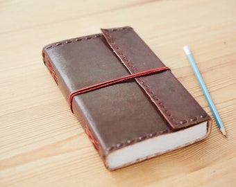 May include: A brown leather-bound journal with a red elastic band closure. The journal is closed and lying on a light wood surface with a blue pencil beside it.
