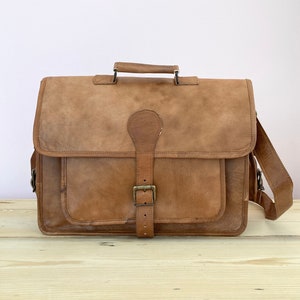 May include: A brown leather briefcase with a buckle closure and a shoulder strap. The bag has a flap with a buckle closure and a handle on the top.