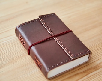 May include: A brown leather-bound journal with a red ribbon closure. The journal has decorative stitching around the edges and a flap that covers the front.