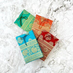 Può includere: Quattro piccoli quaderni con copertine decorative. Le copertine sono realizzate in tessuto con motivi intricati in tonalità di blu, arancione e rosso. Ogni quaderno ha un nastro verde legato intorno.
