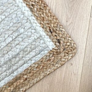 May include: A close-up of a jute rug with a white border. The rug is woven with a herringbone pattern and has a natural, earthy tone.