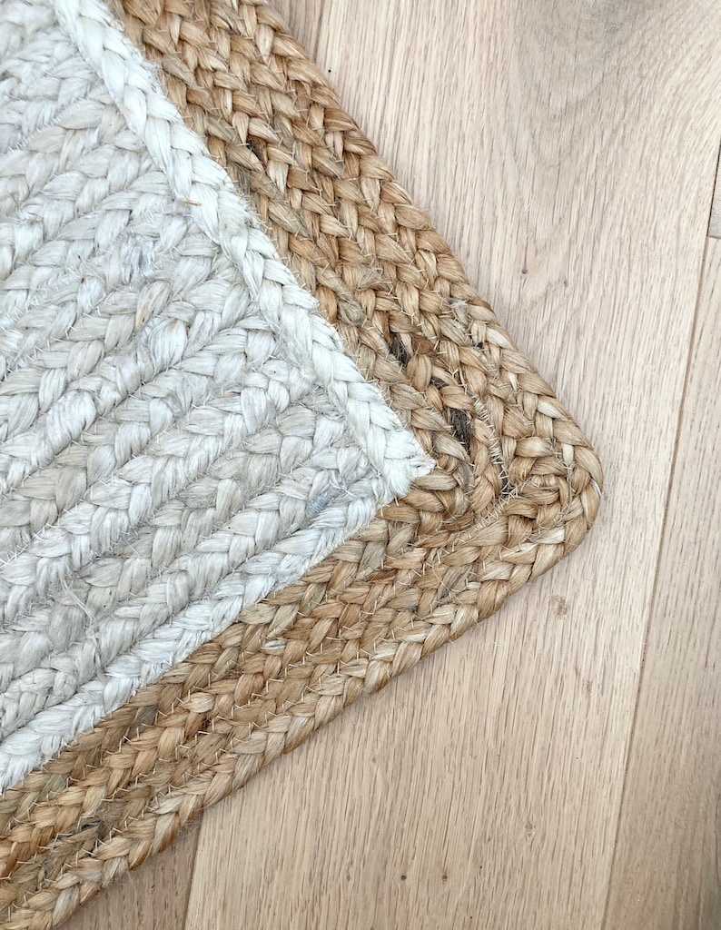 May include: A close-up of a jute rug with a white border. The rug is woven with a herringbone pattern and has a natural, earthy tone.