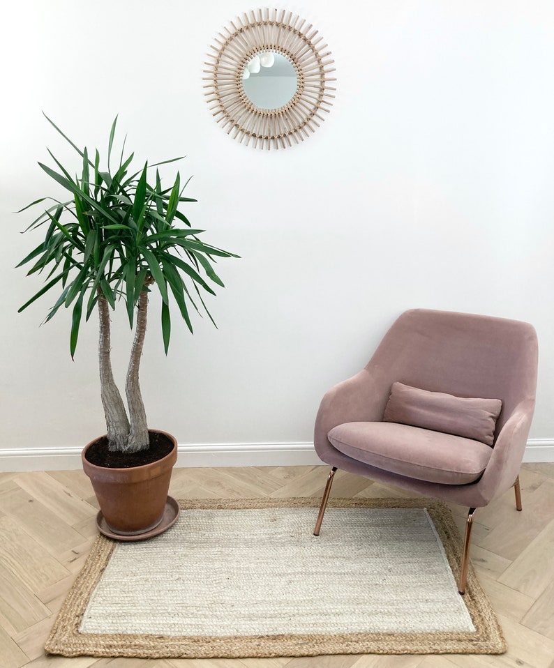 May include: A pink velvet armchair with a pillow sits on a light beige rug with a jute border. A potted green plant and a round woven mirror with a white background are on the wall behind the chair.