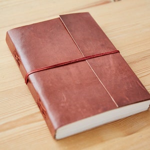 Peut inclure: Un journal relié en cuir marron avec une fermeture en ruban rouge. Le journal est fermé et posé sur une surface en bois.