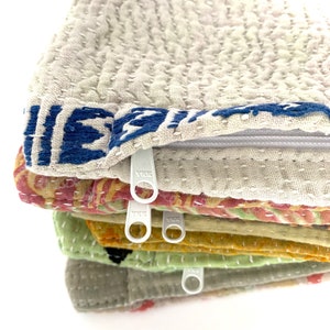 May include: A stack of six fabric zipper pouches in various colors and patterns, including beige, green, yellow, and pink. The pouches are made of a soft, quilted fabric and have white zipper pulls.