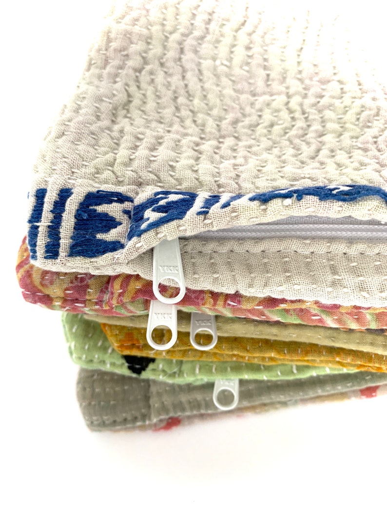 May include: A stack of six fabric zipper pouches in various colors and patterns, including beige, green, yellow, and pink. The pouches are made of a soft, quilted fabric and have white zipper pulls.