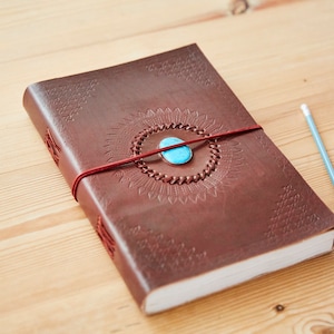 May include: A brown leather journal with an intricate design and a turquoise stone accent. The journal is closed and tied with a red string.