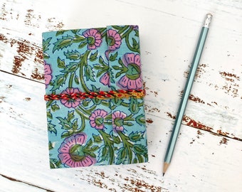 May include: A small blue notebook with a floral pattern and a red and gold braided cord closure. The notebook is lying on a white wooden surface with a pencil beside it.