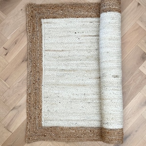 May include: A white jute rug with a brown jute border. The rug is rolled up and ready to be placed on the floor.