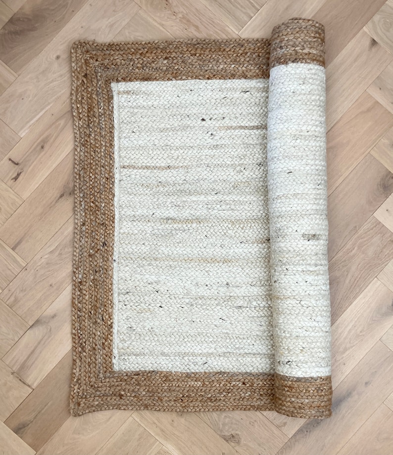 May include: A white jute rug with a brown jute border. The rug is rolled up and ready to be placed on the floor.