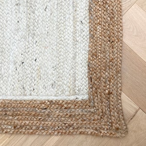 May include: Close-up of a white and natural jute rug with a thick, braided border. The rug is woven with a simple, textured pattern.