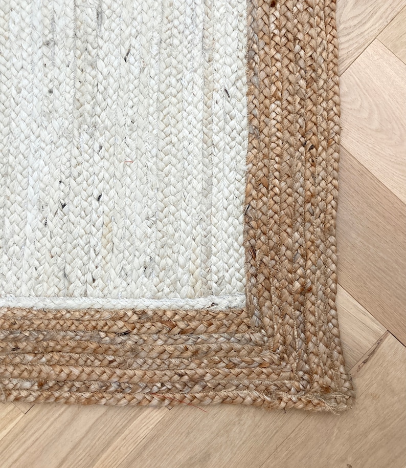 May include: Close-up of a white and natural jute rug with a thick, braided border. The rug is woven with a simple, textured pattern.