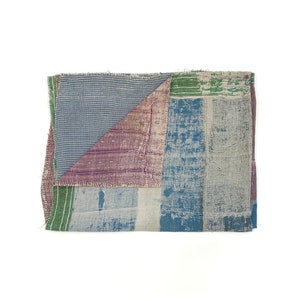 Op de afbeelding: Een patchworkdeken gemaakt van hergebruikt textiel in tinten blauw, groen en paars. De deken is dubbelgevouwen en heeft een ruw, getextureerd oppervlak.