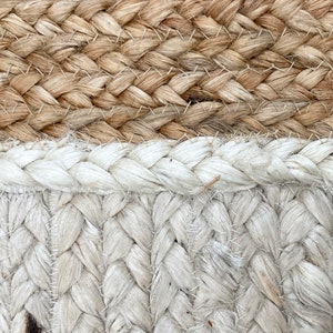 May include: Close-up of a woven rug with a natural brown and white color scheme. The rug is made of thick, braided rope and has a textured surface.