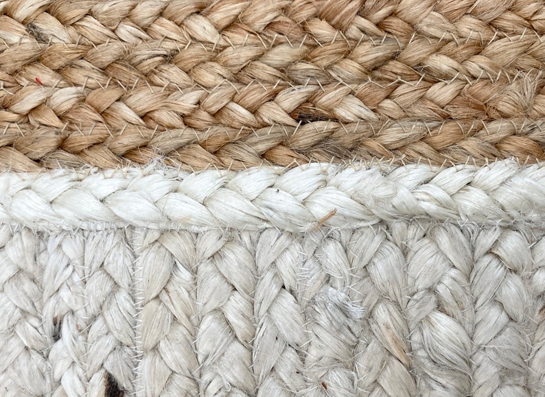 May include: Close-up of a woven rug with a natural brown and white color scheme. The rug is made of thick, braided rope and has a textured surface.
